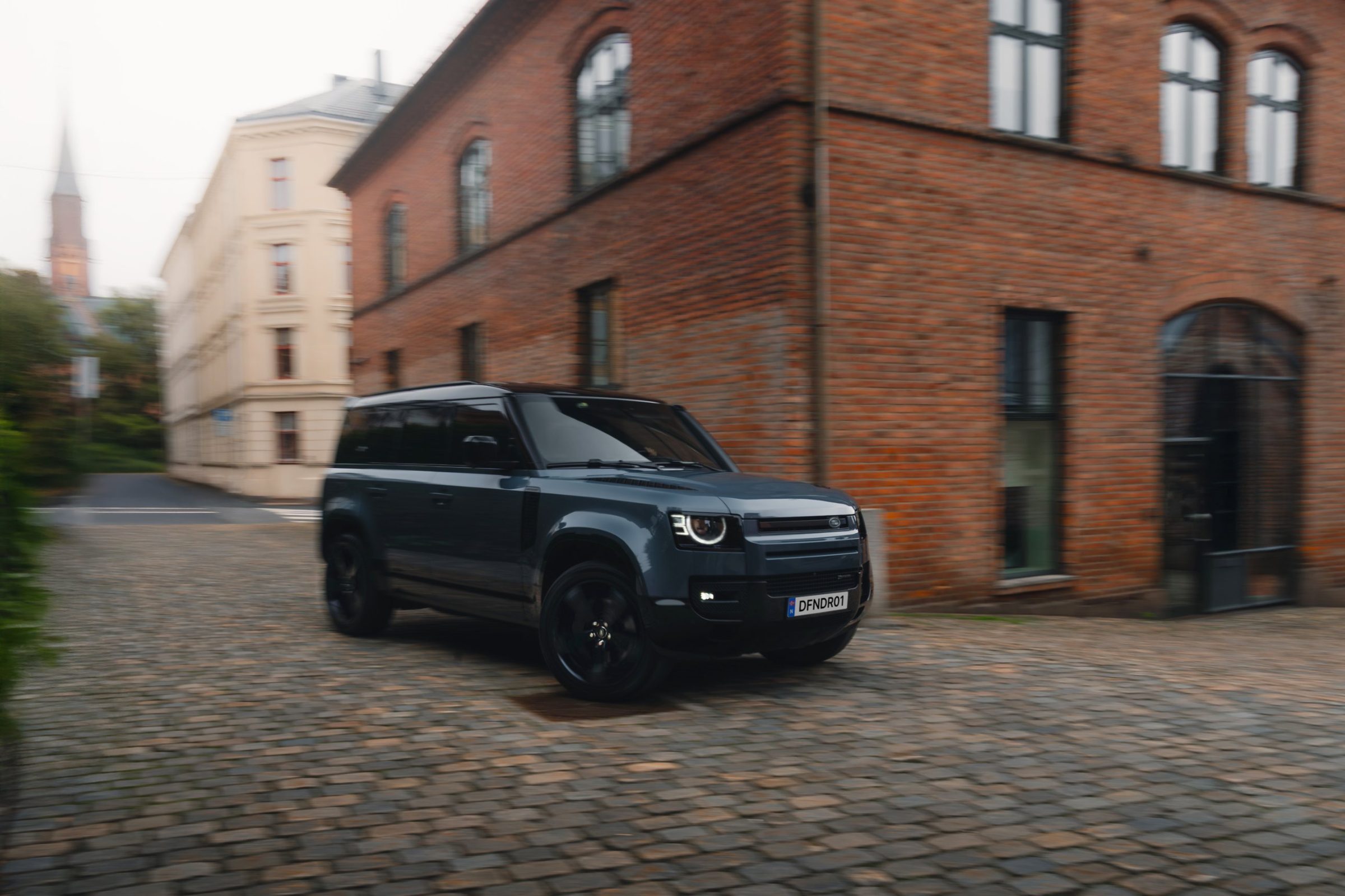 Blå Land Rover Defender i fart over brostein foran et gammelt teglsteinsbygg